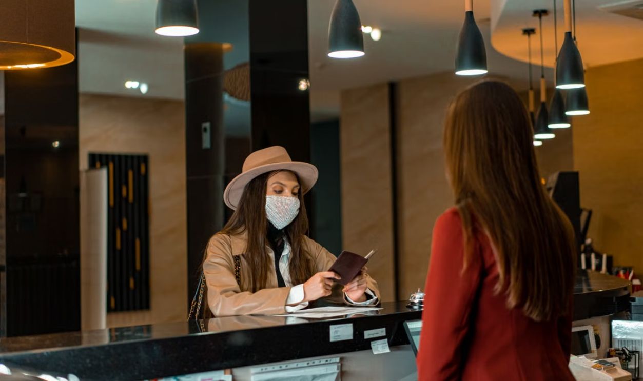 A woman wearing a mask checks in at a hotel reception desk.