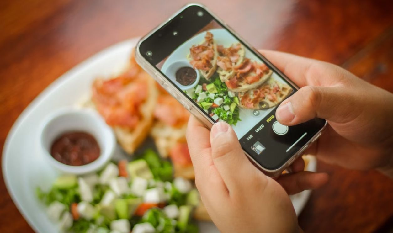 Close-up of hands photographing a brunch plate with bacon, salad, and sauce using a smartphone
