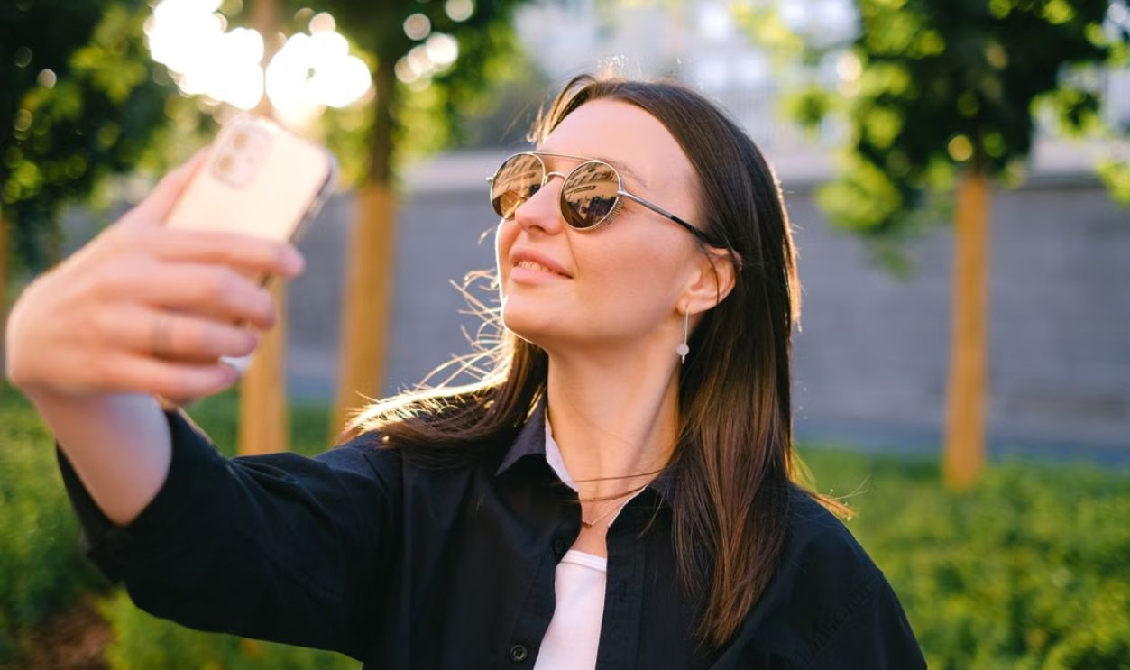 Smiling woman in sunglasses takes selfie outdoors. Bright day, casual style, smartphone camera
