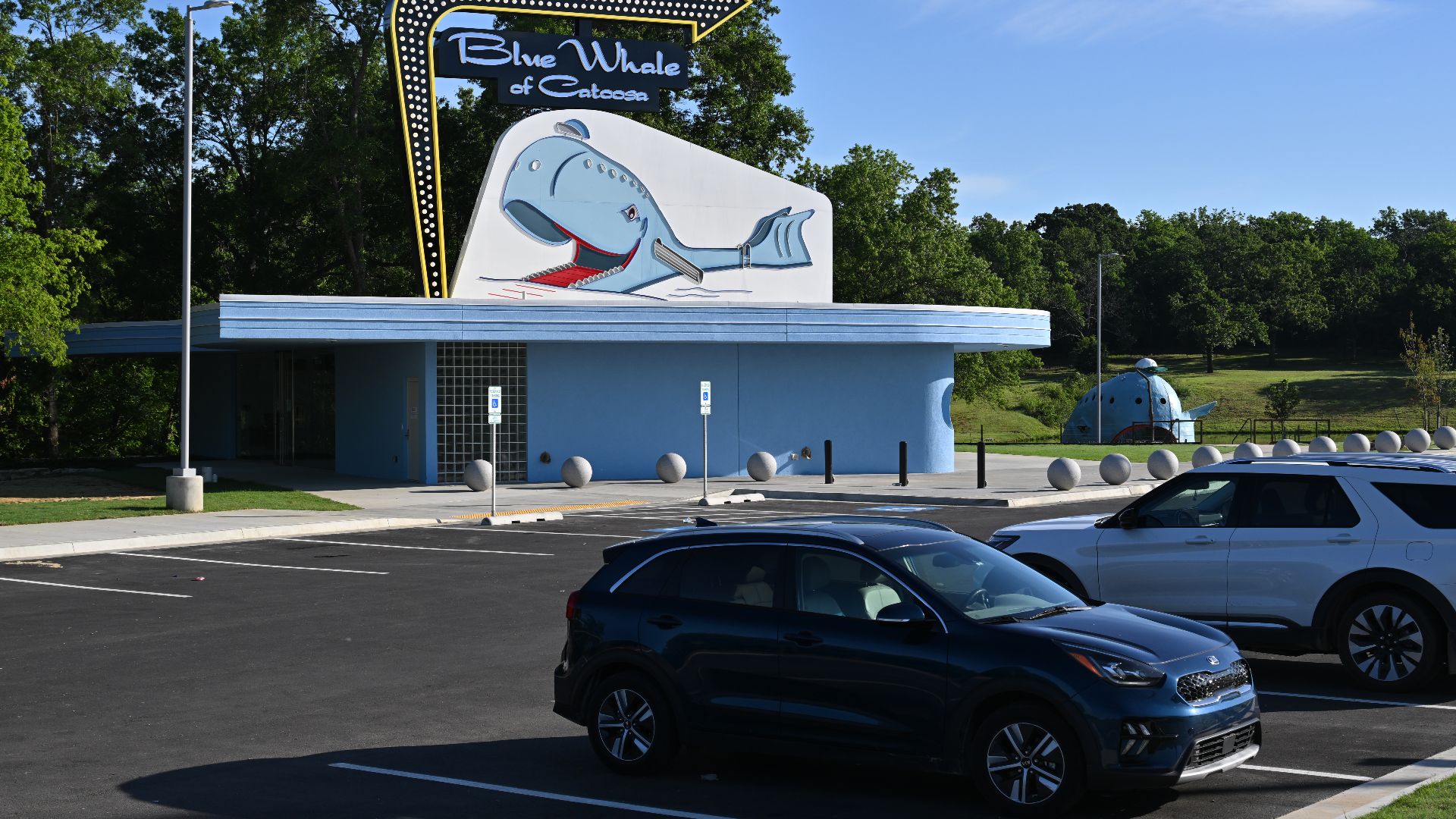 The Blue Whale of Catoosa visitor center with a large sign above it. To the right is the actual Blue Whale and in front of the visitor center is a freshly paved parking lot with cars parked in it.  This photo was taken on Monday May 25, 2026, but the visi