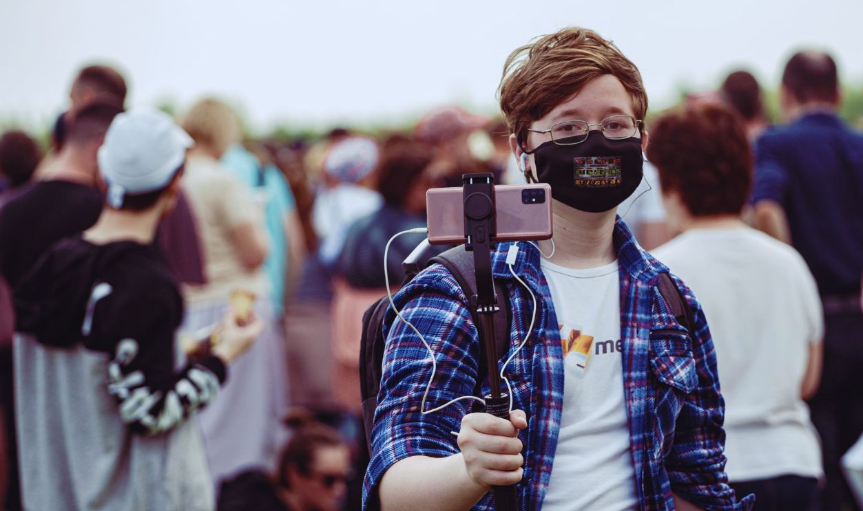 Young individual wearing a mask records a crowd with a smartphone mounted on a selfie stick