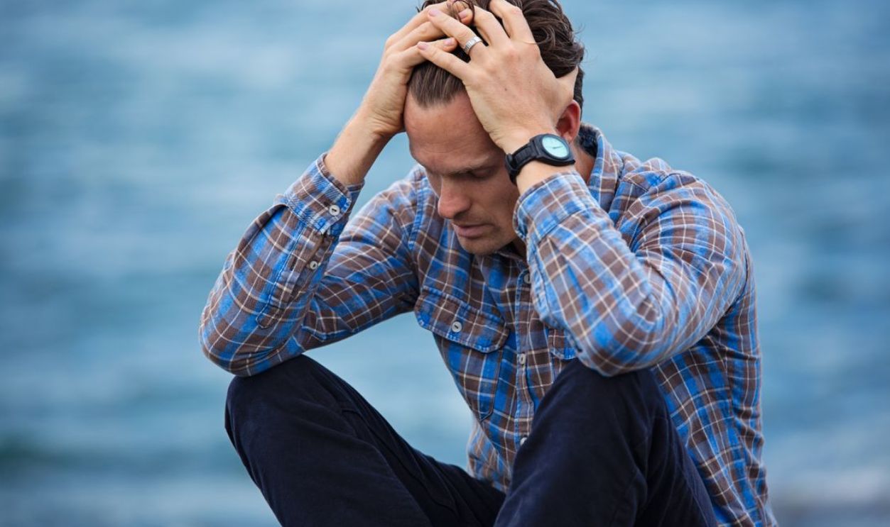 A man in a plaid shirt sits by the water looking distressed, symbolizing stress