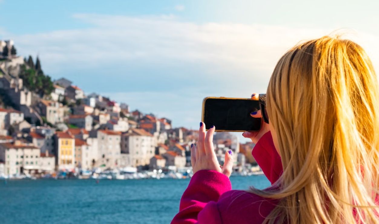 A woman takes a photo of the Sibenik waterfront using a smartphone on a sunny day
