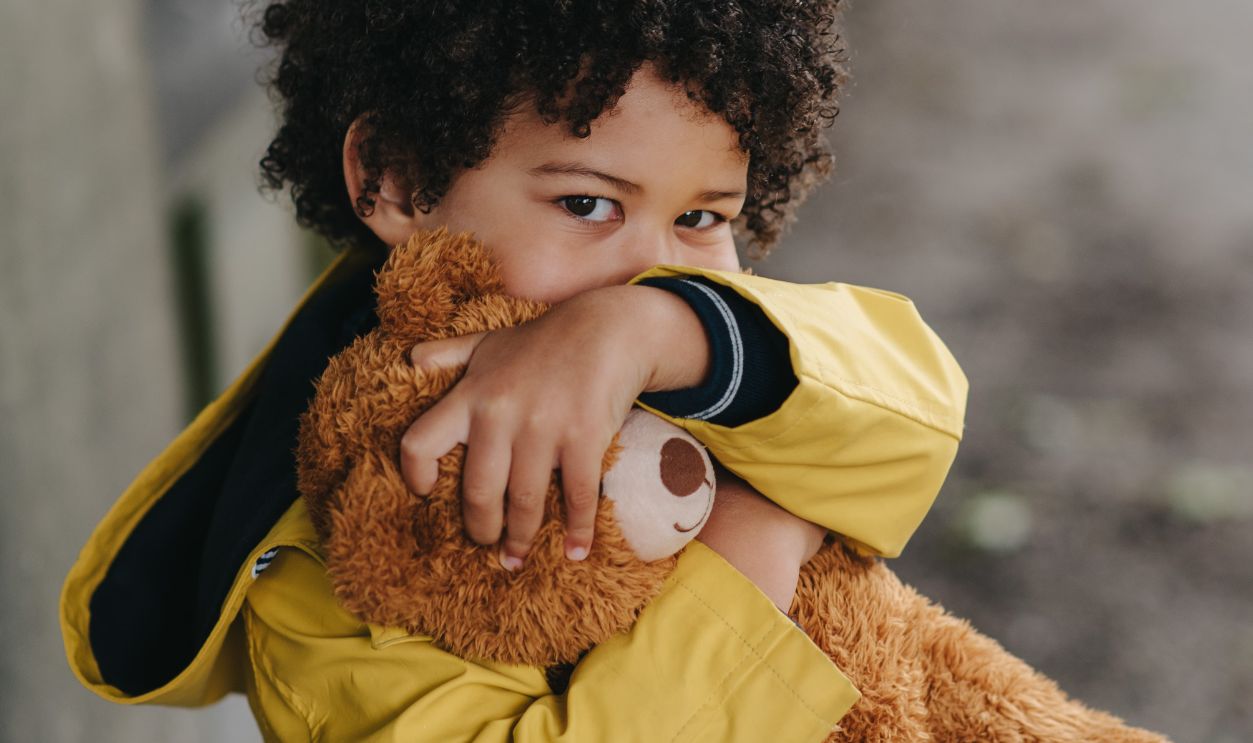 A cute child in a yellow jacket hugs a teddy bear with a blurred background.