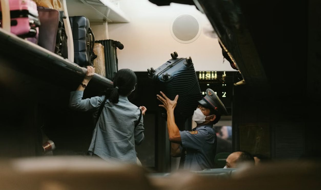 Train attendant helps passenger with luggage in overhead compartment during travel.