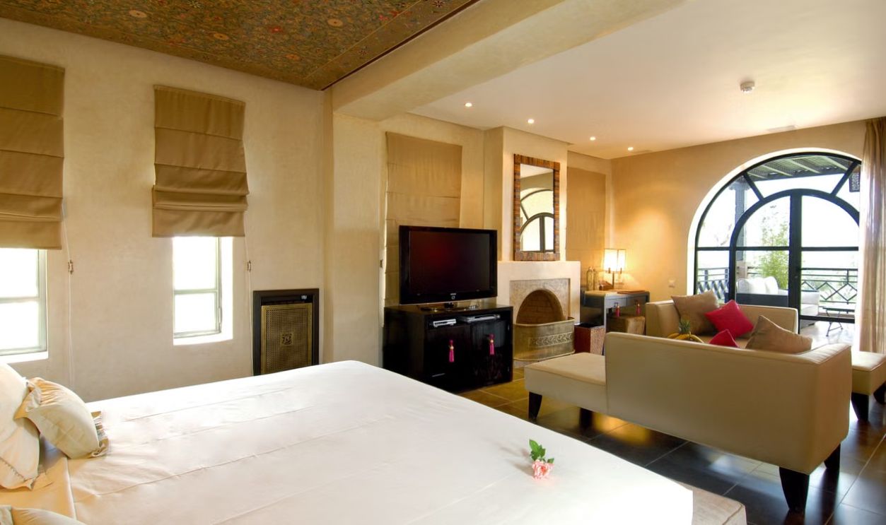 Elegant modern bedroom suite with cozy seating, large windows, TV, and fireplace, exuding warmth and luxury.