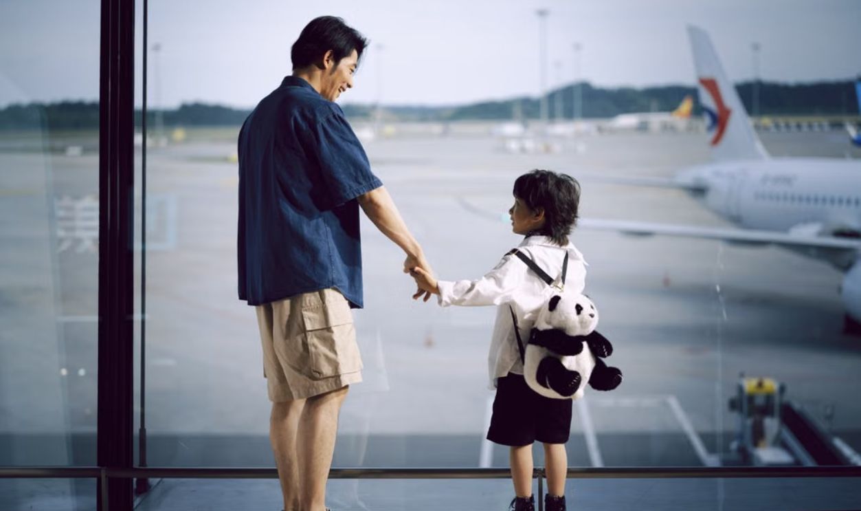 A father and child holding hands, gazing at planes through an airport window.