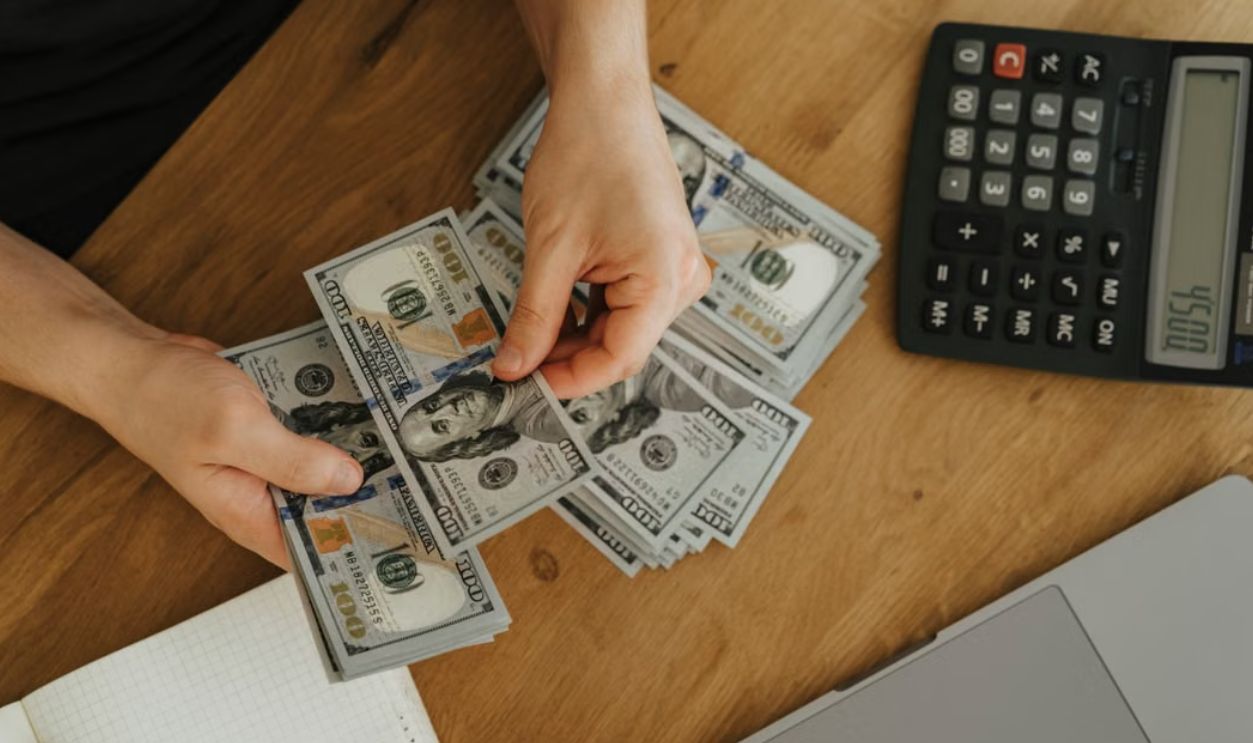 Close-up of hands counting dollar bills with calculator, notebook, and laptop on wooden table