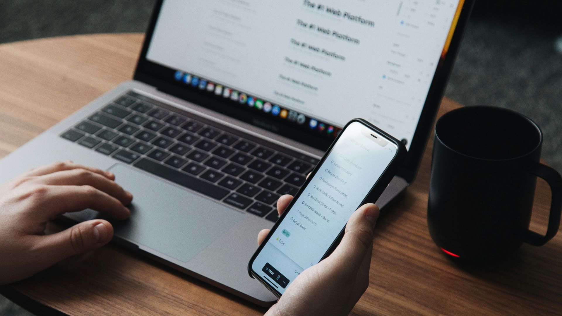 person holding smartphone in front of turned-on laptop beside black ceramic mug on oval brown wooden table