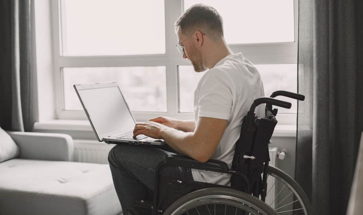 A man in a wheelchair is using a laptop in a bright room with a large window.
