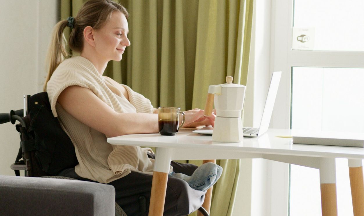 A woman in a wheelchair working on a laptop in a bright indoor space with green curtains