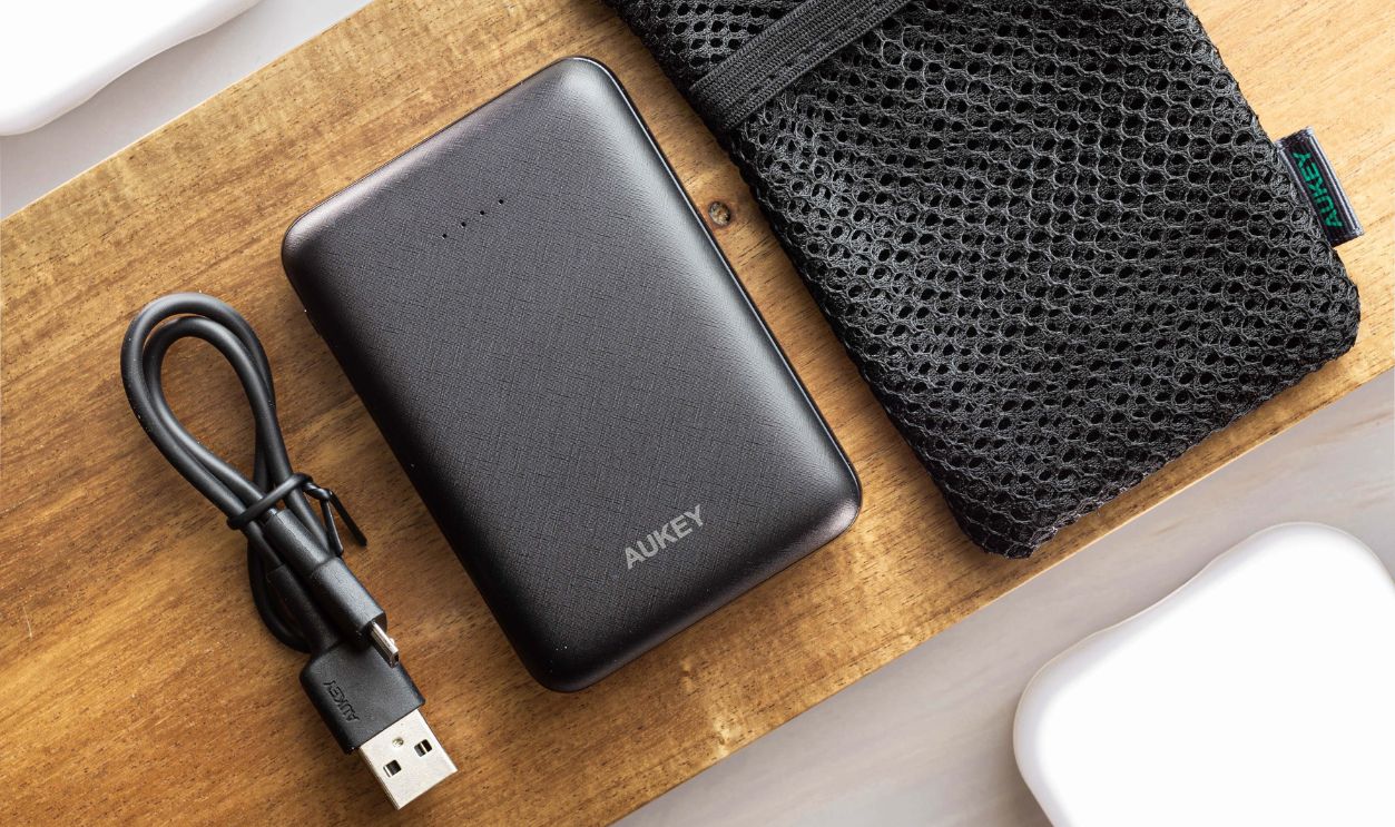 Flat lay of a black power bank with a USB cable and mesh pouch on wood.