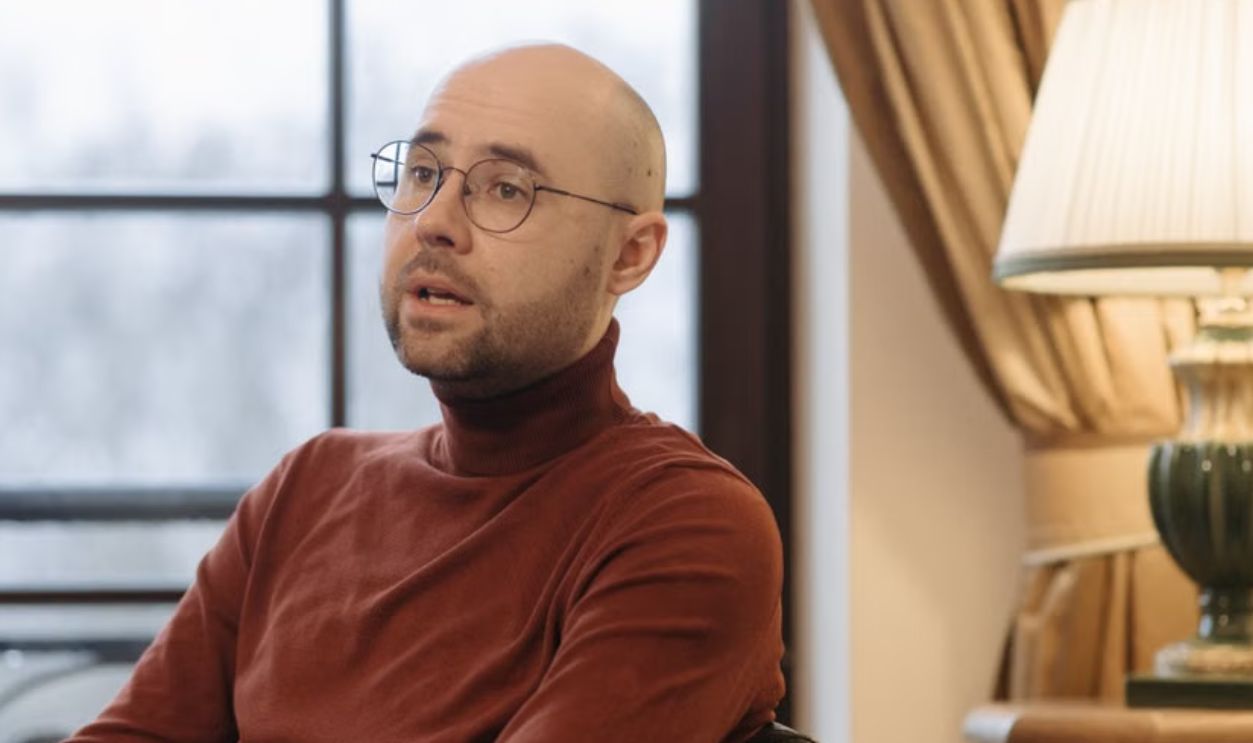 Bald man in glasses speaking while seated indoors, with a warm lamp and curtains in the background
