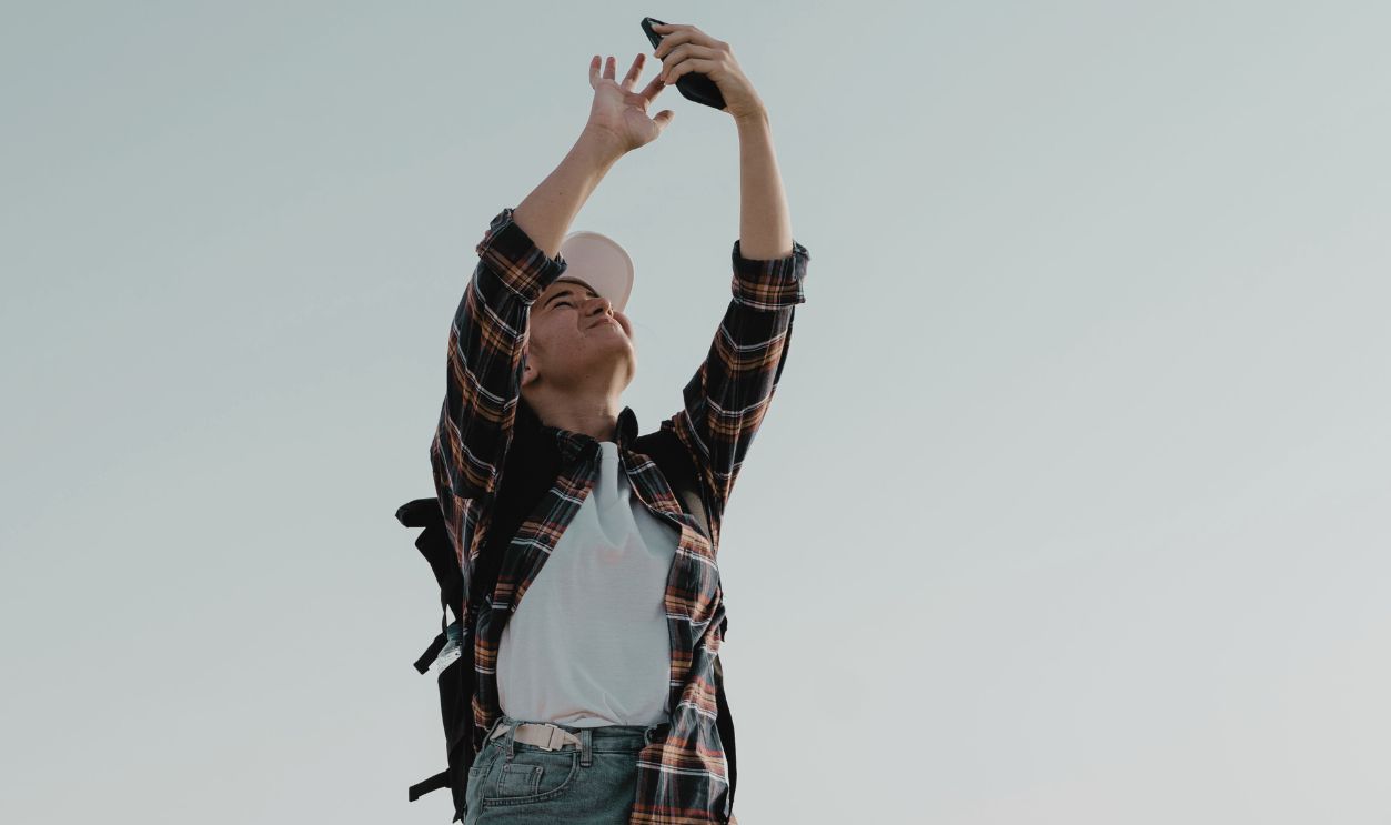 A young woman in casual attire taking a selfie against a clear sky.