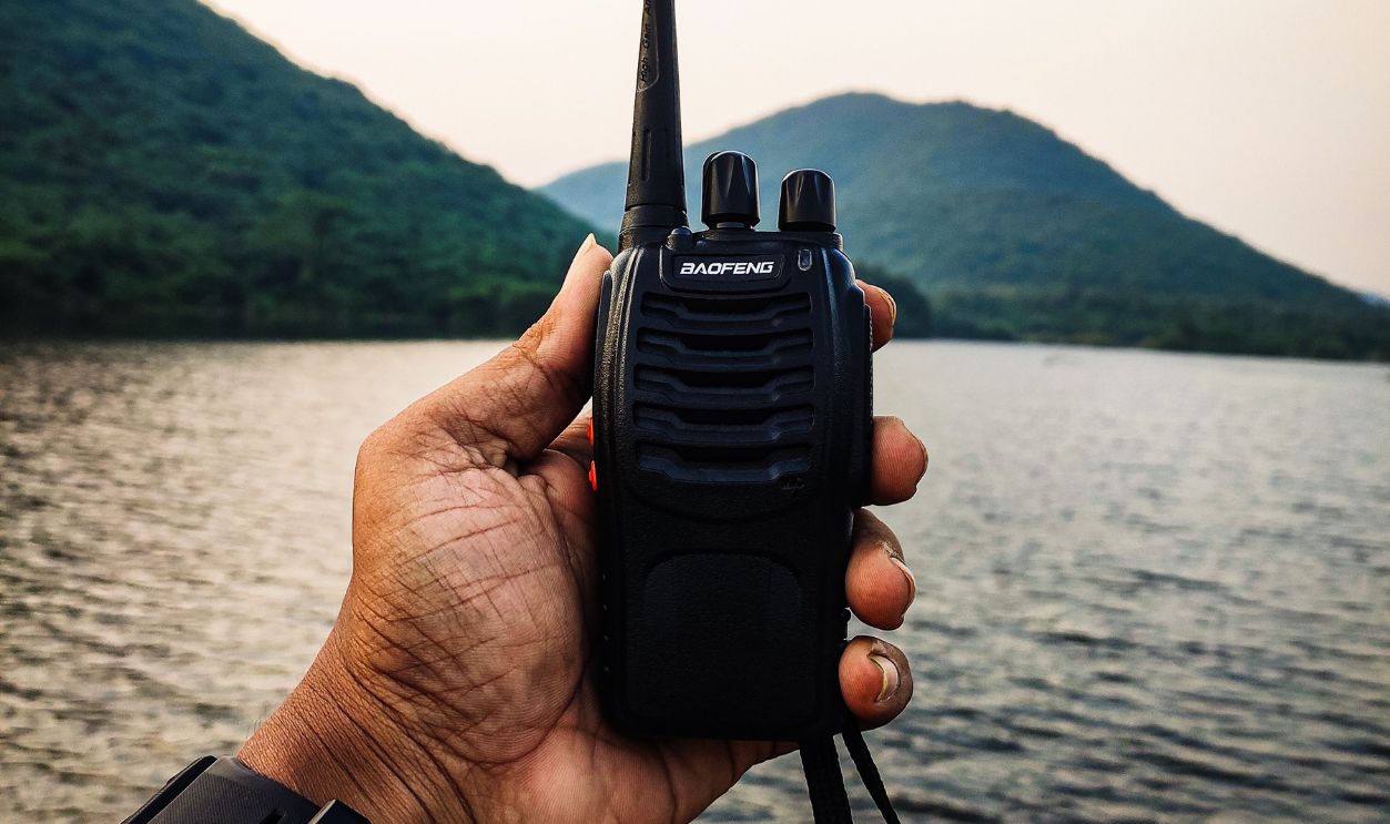 Close-up of a hand holding a walkie talkie with a lake and mountains in the background.