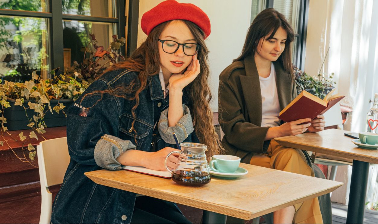 Two women relaxing at a café, one reading a book and the other wearing a beret, sipping coffee
