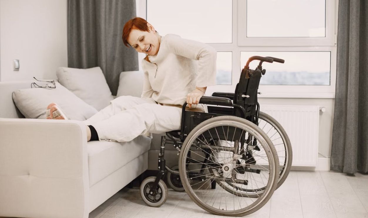 A woman with dyed short hair smiles while transitioning from a wheelchair to a couch