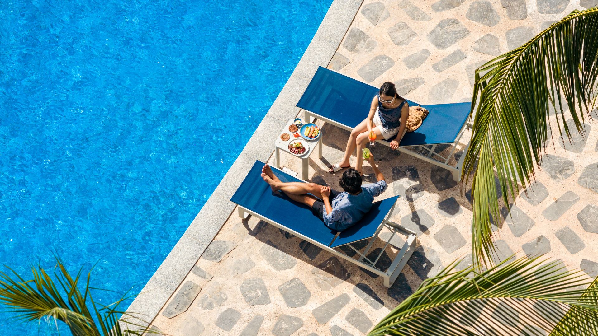 Two people relaxing on sun loungers by a blue pool.