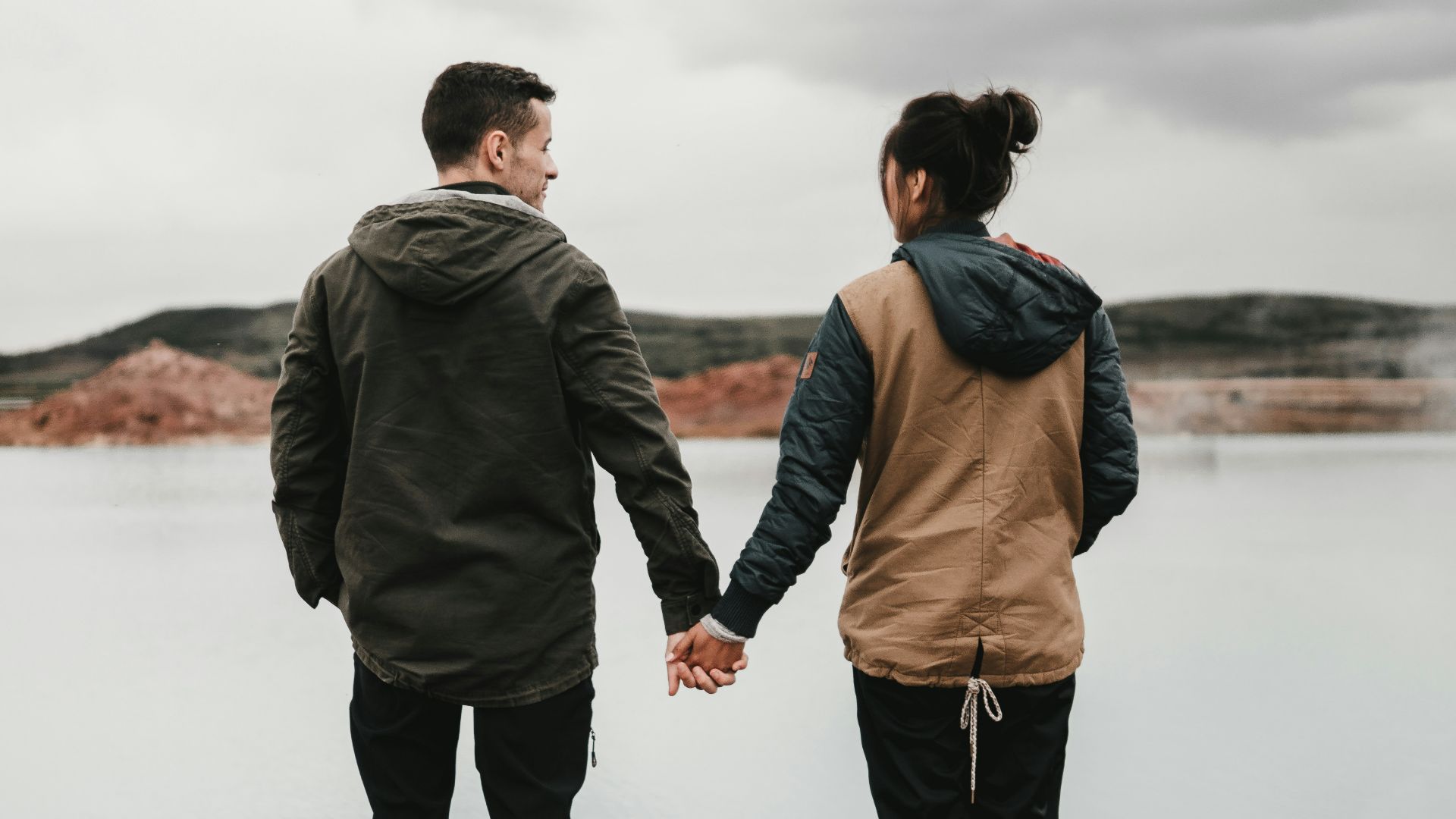 couple holding hand front of body of calm water with mountain distance