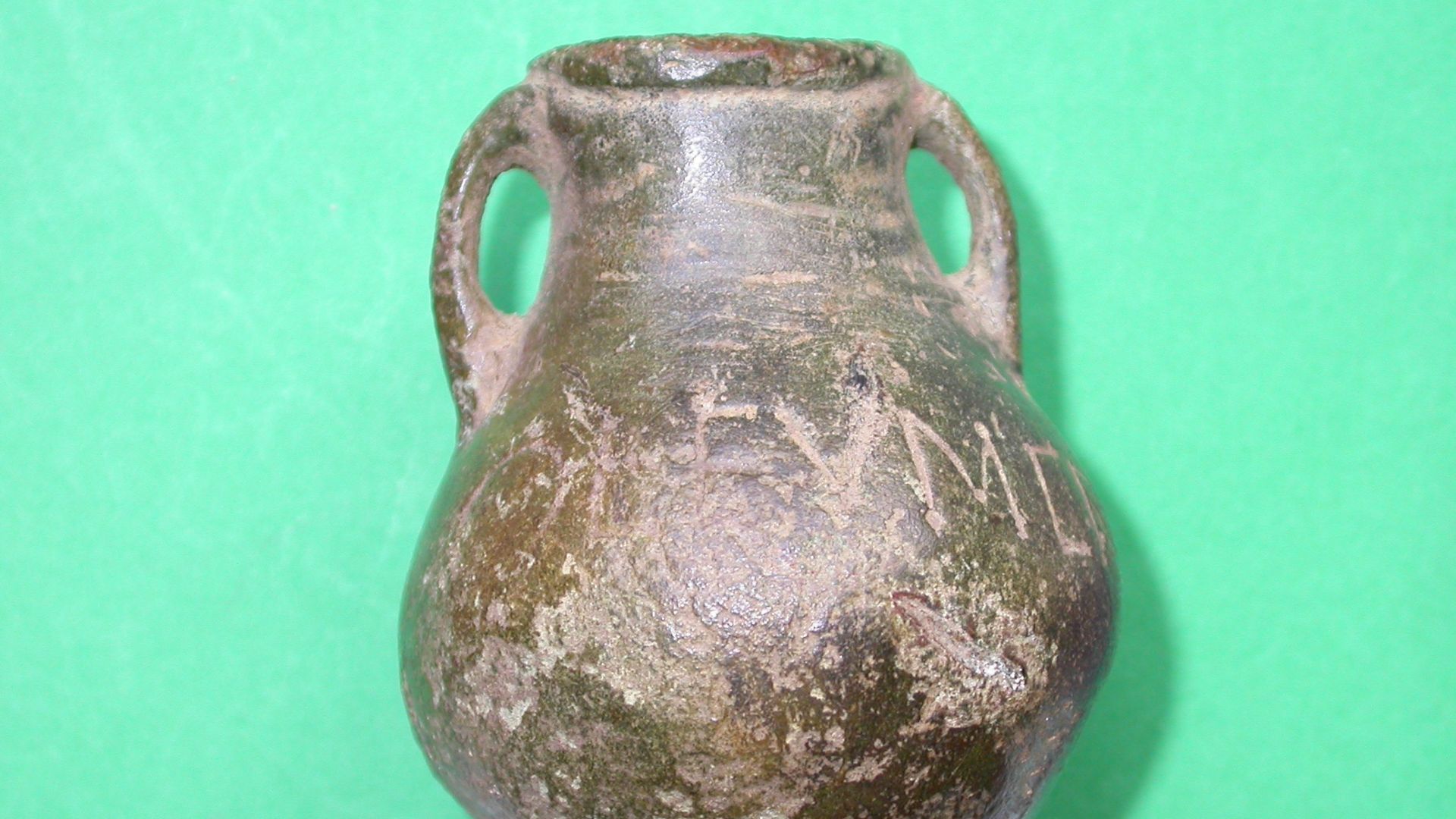 A miniature two-handled vessel with rounded base and beaded rim, cast in one piece. The vessel's Latin inscription (OLEVM CHRISM) is crudely applied, and the final M is a little unclear due to wear.. The terminals and the junctions of some of the letters 