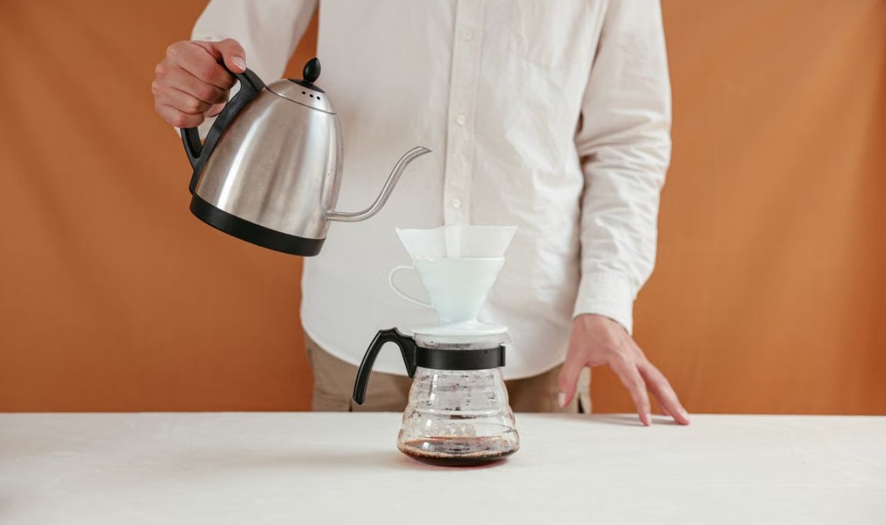 A person brewing pour over coffee using a dripper and kettle in a minimalistic setting.