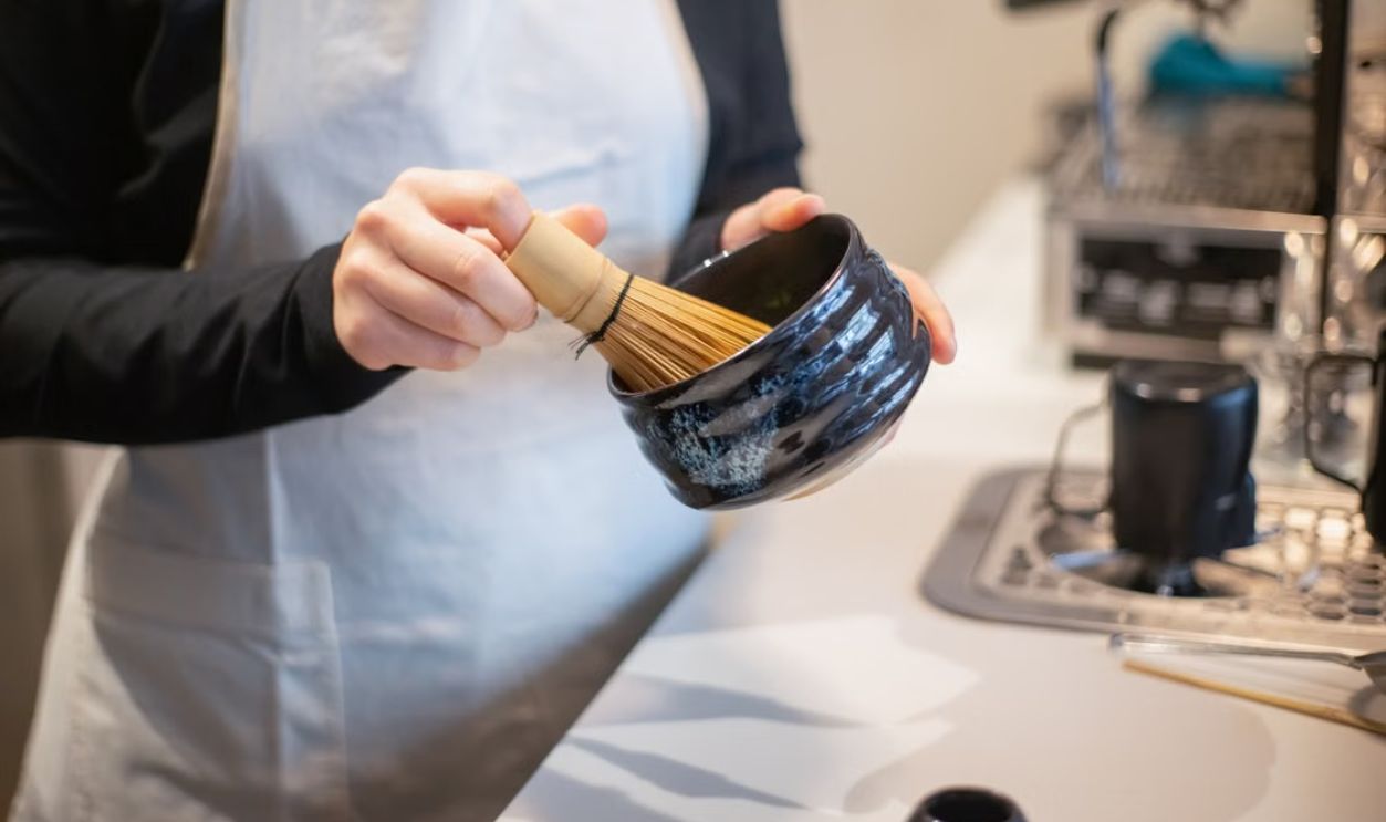 Close-up of a person preparing matcha tea using a bamboo whisk and bowl indoors.