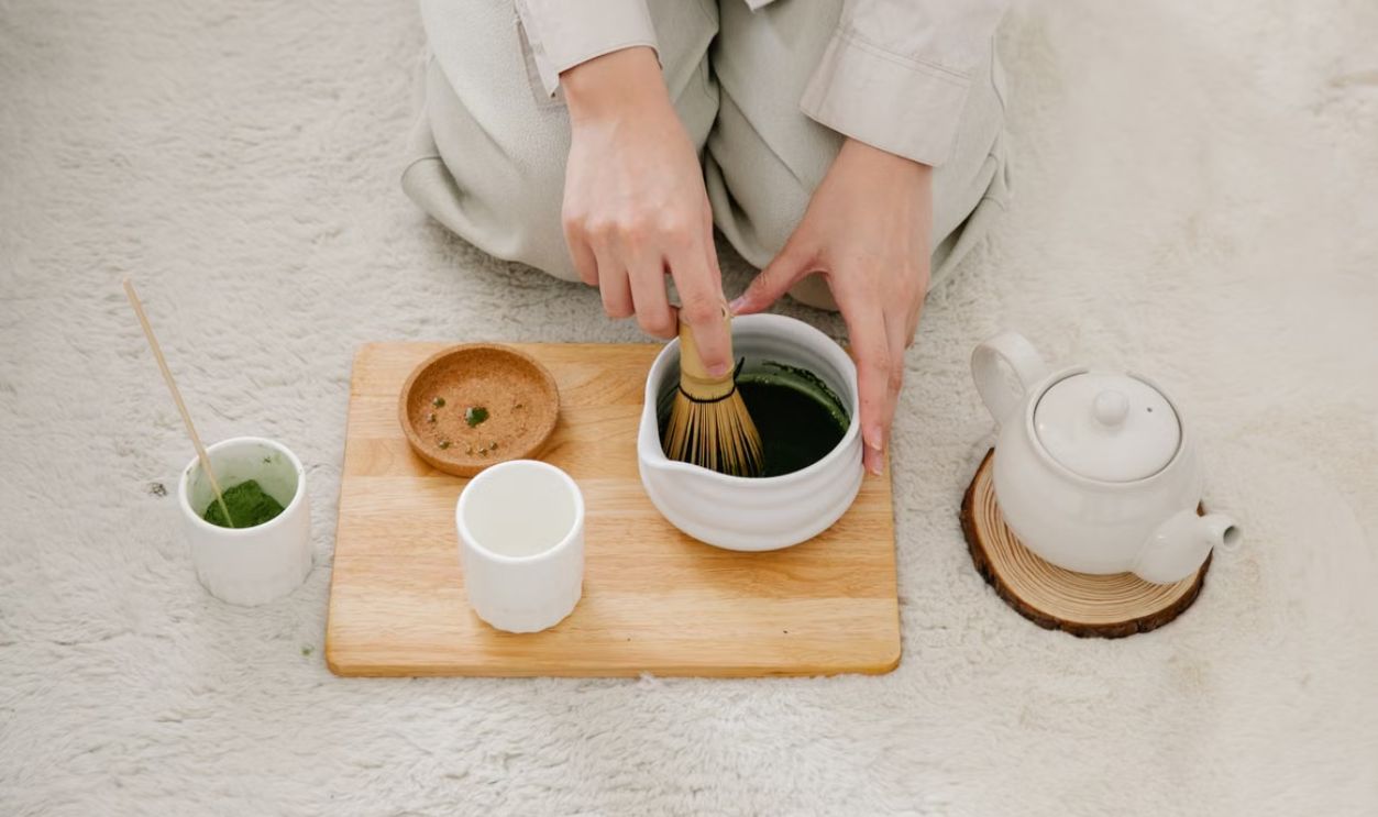 Hands preparing matcha tea with a whisk on a wooden board. Includes teapot and cups.