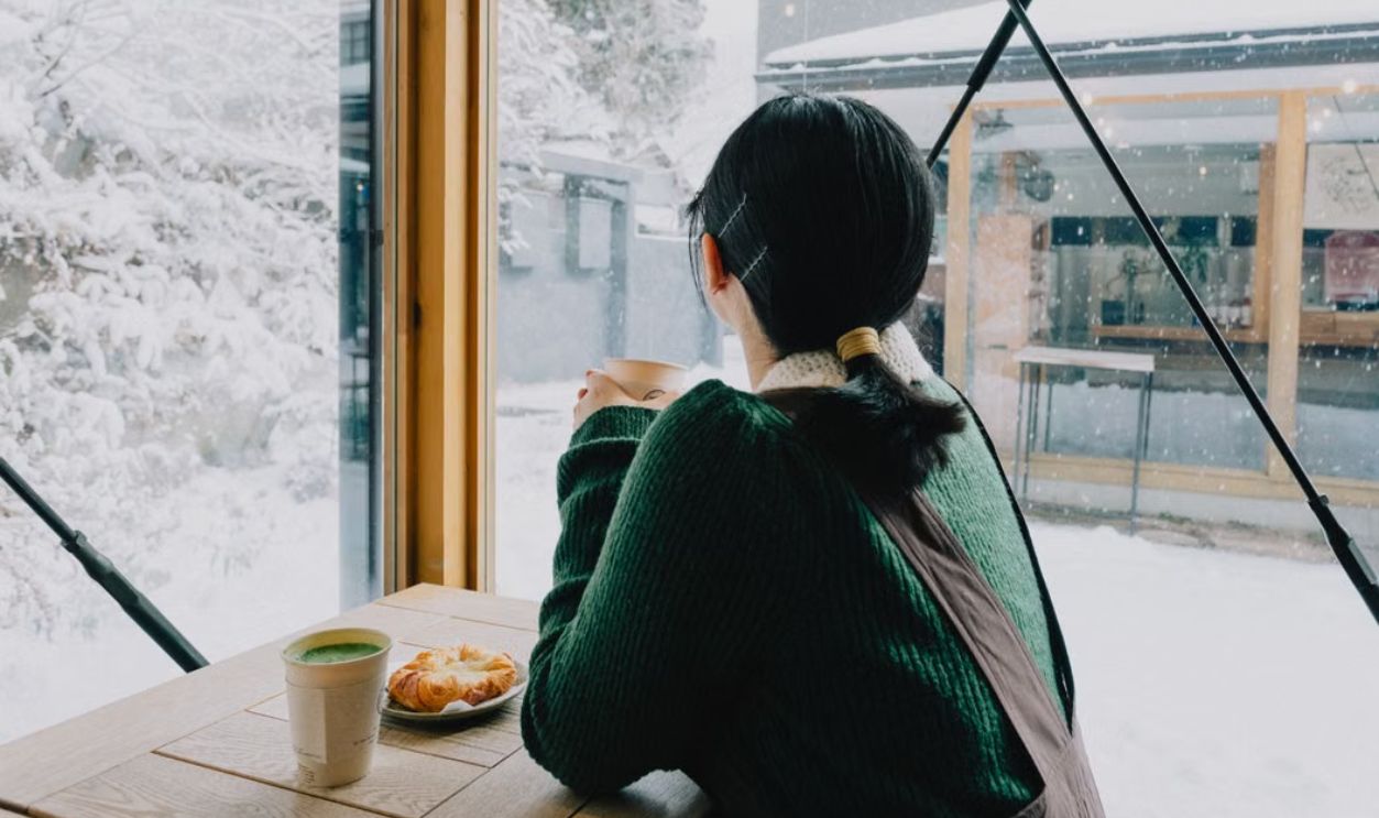 Woman enjoying coffee inside a cafe, gazing at snow-covered landscape in Nikko, Japan.