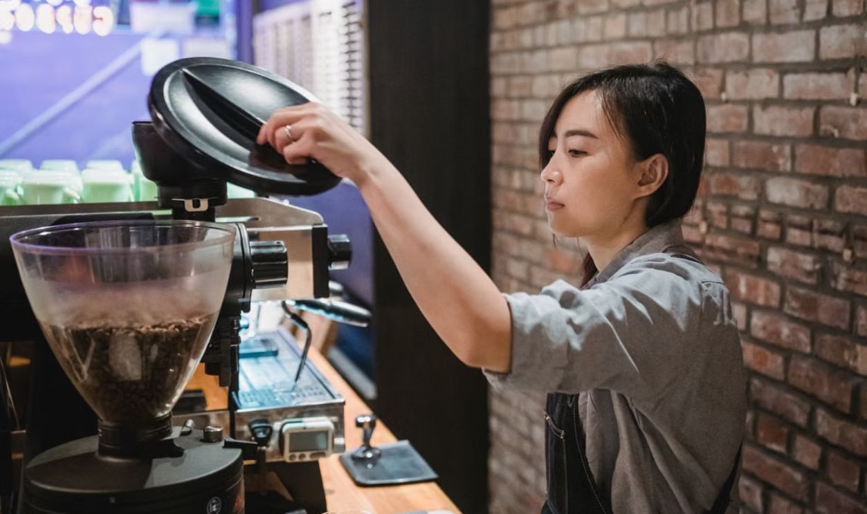 A focused barista prepares coffee using a grinder in a cozy, brick-walled cafe setting.