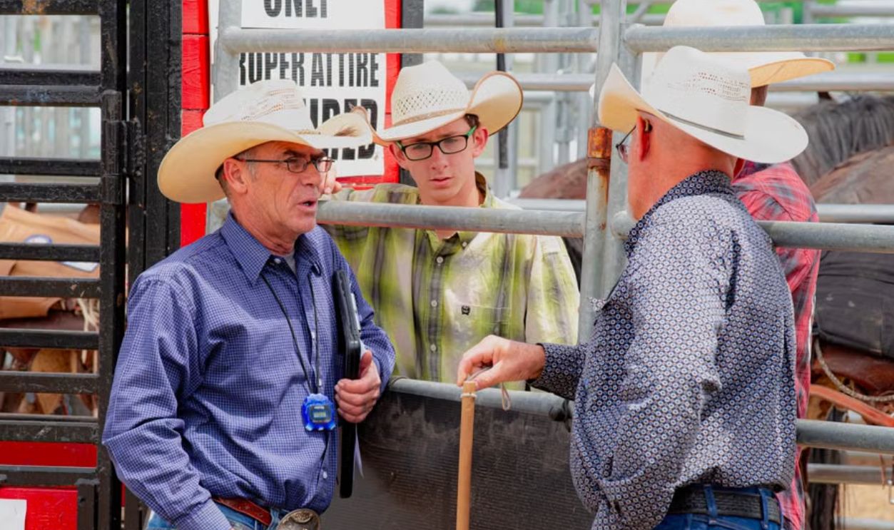 Three cowboys in conversation at a rodeo event gate, wearing iconic hats and attire