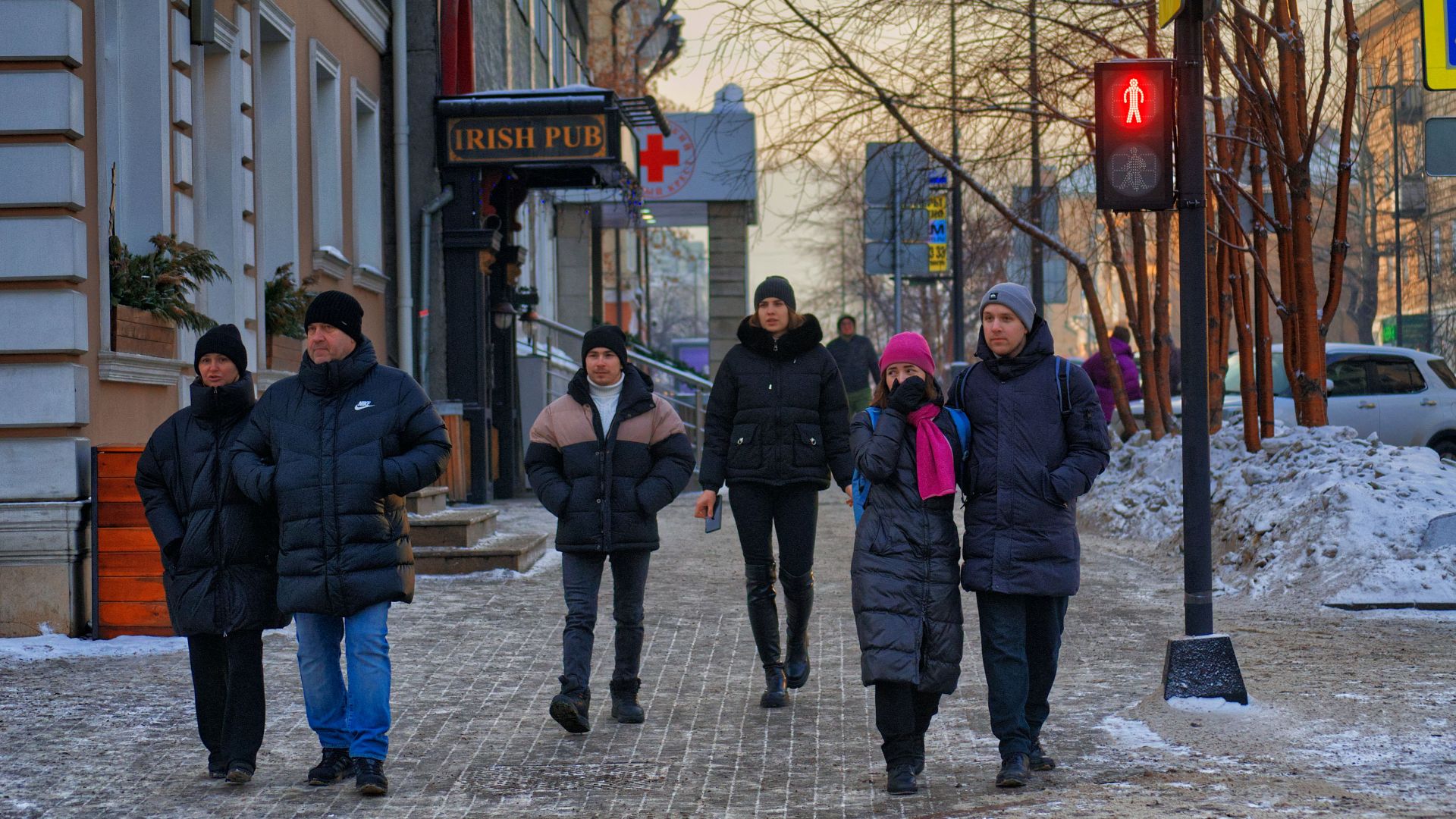 People walking on a snowy sidewalk in a city during winter with a red pedestrian light.