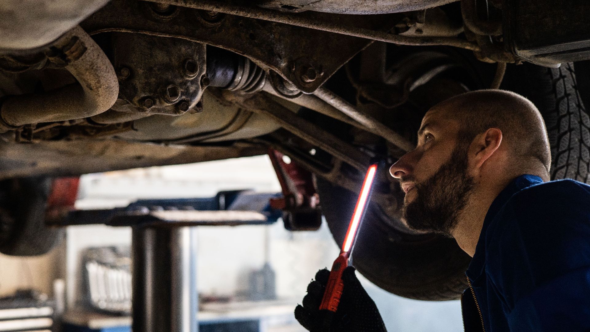 Mechanic using light to inspect vehicle undercarriage in workshop for maintenance.