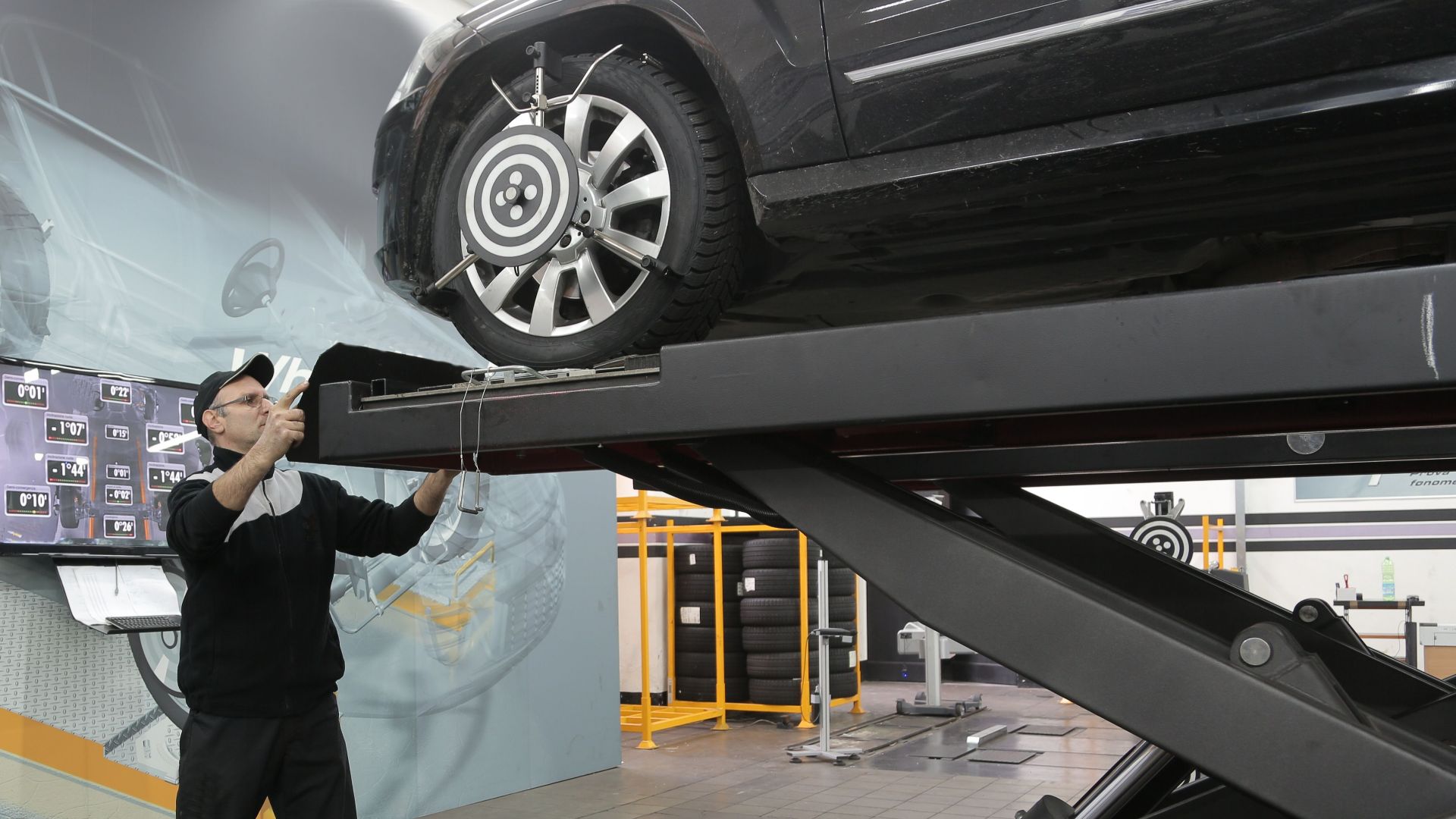 Serious professional adult mechanic in casual uniform examining car wheels while standing near lift in modern car service garage
