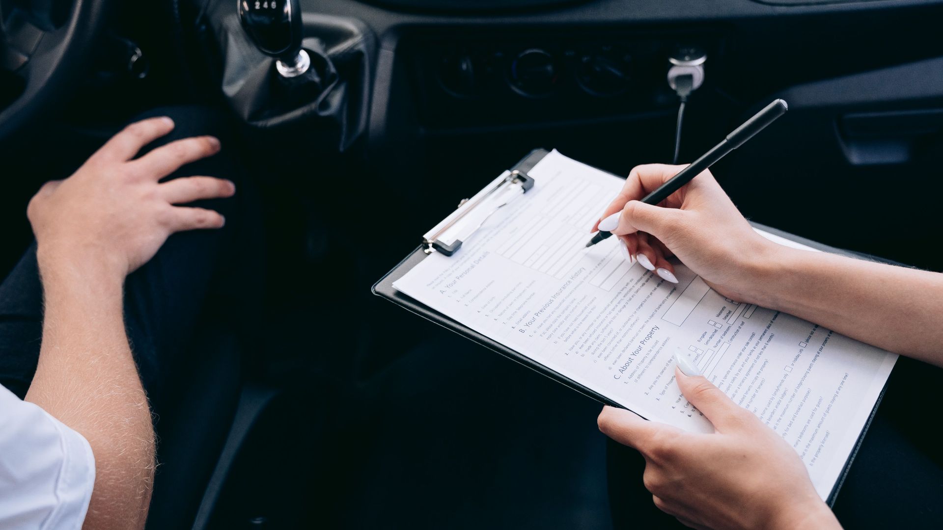 Close-up of a person writing on a clipboard inside a car, showing hands and a gear shift.