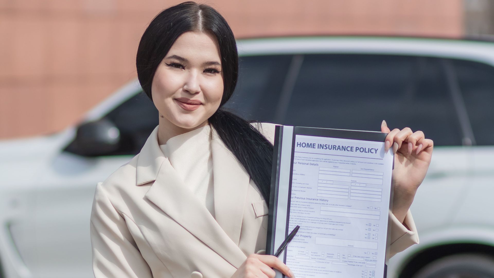 Smiling woman in beige blazer holding a home insurance policy document in outdoor setting.