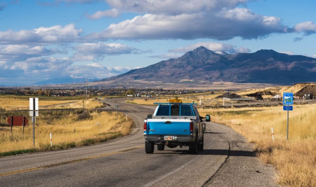 A blue pickup truck drives on a rural road with scenic mountain views under a blue sky