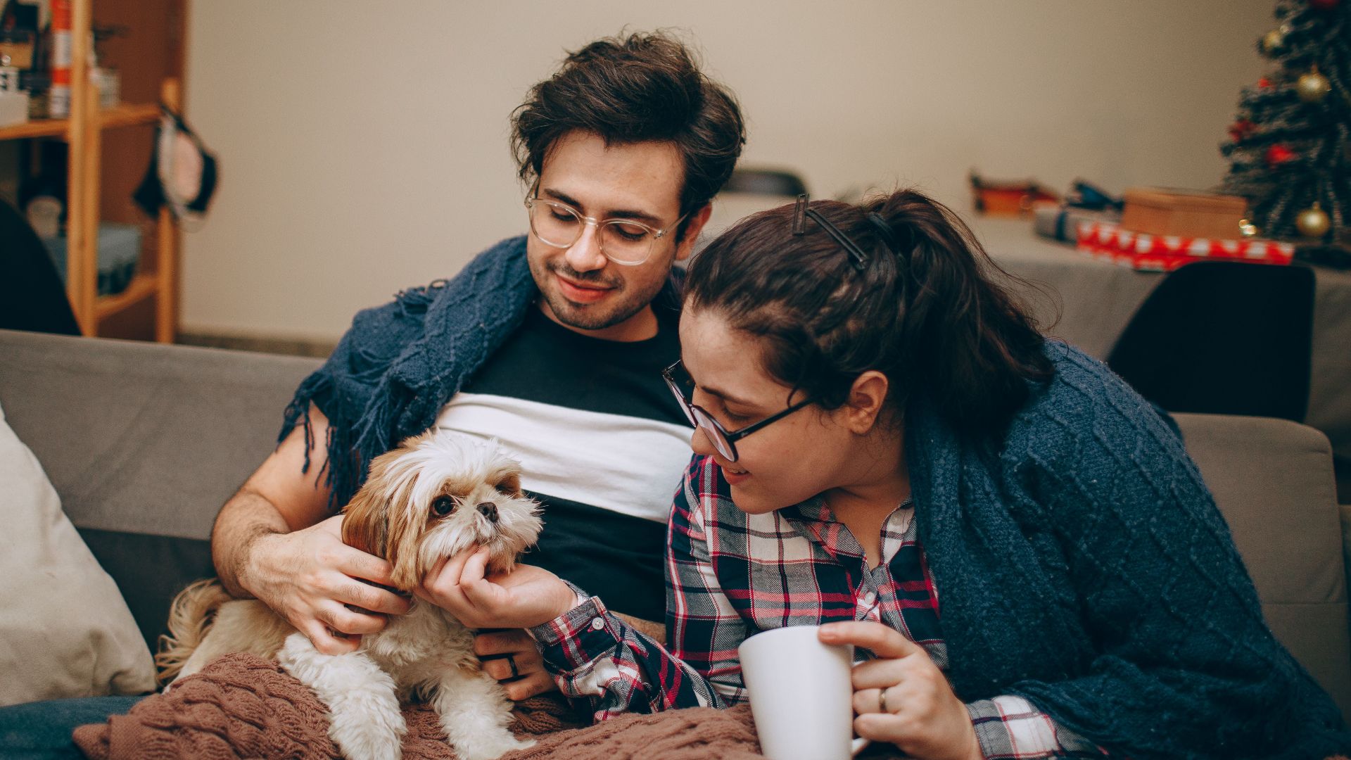 Happy couple relaxing indoors with their cute pet dog during winter, wrapped in blankets.