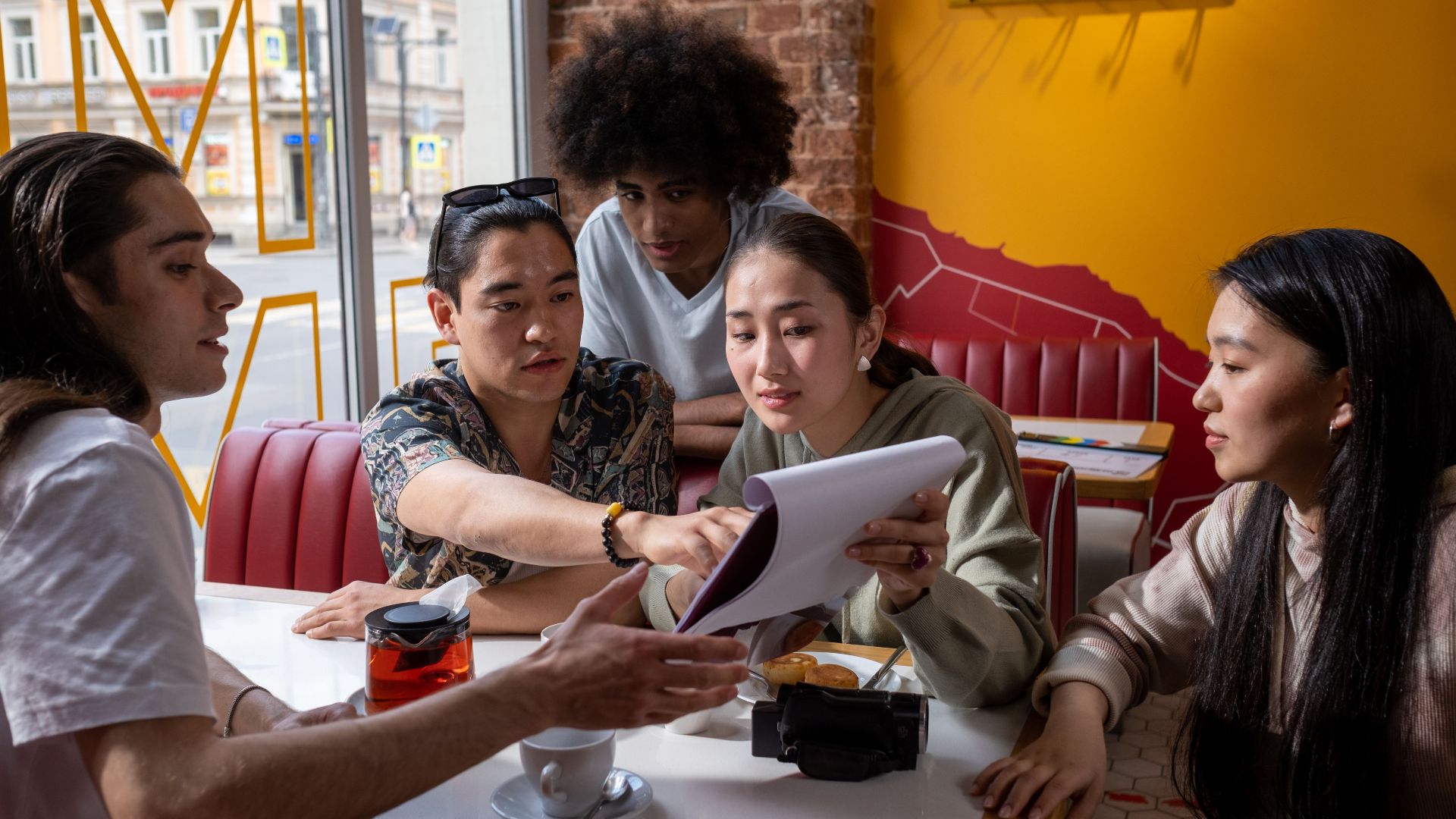 A multicultural group of young adults engaged in conversation around a table in a cafe, focused on a document.