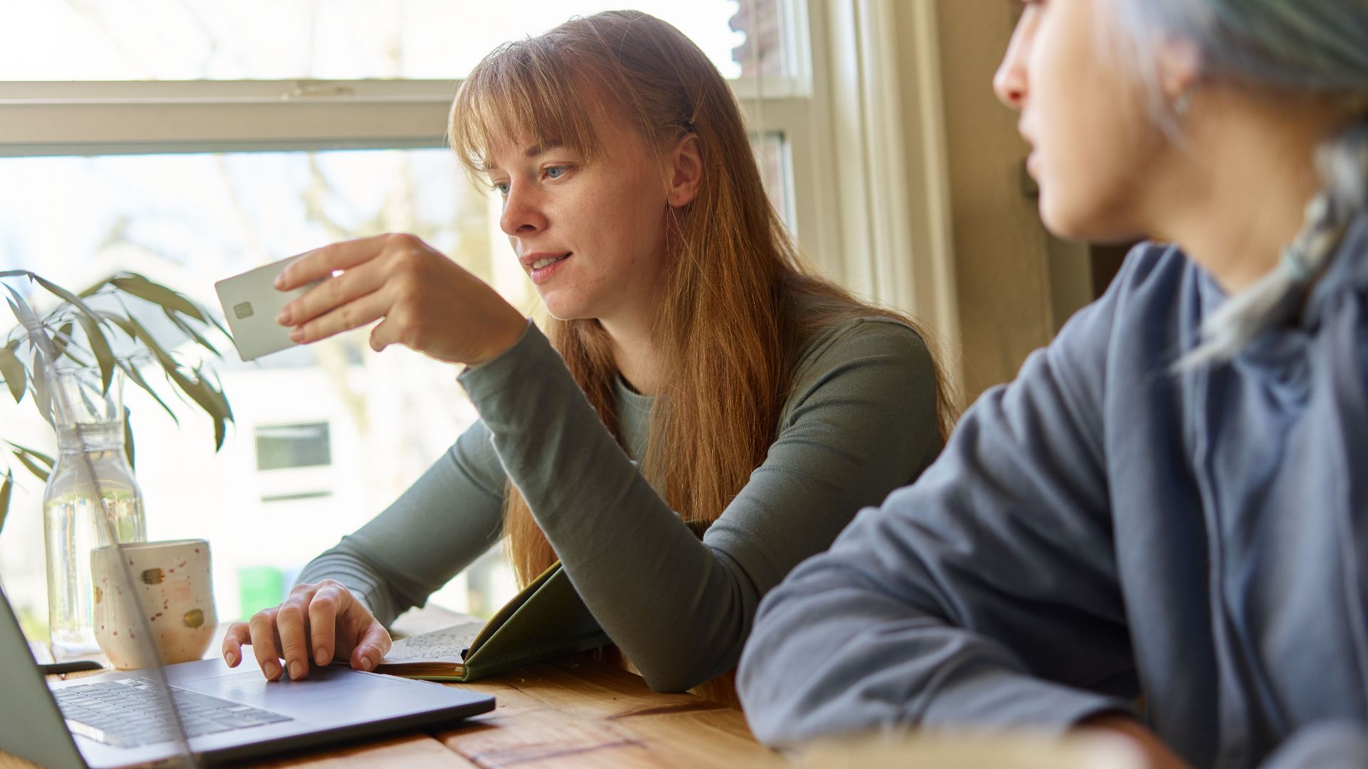 Two young women in casual attire discussing online payments using a laptop at a wooden table near a window.
