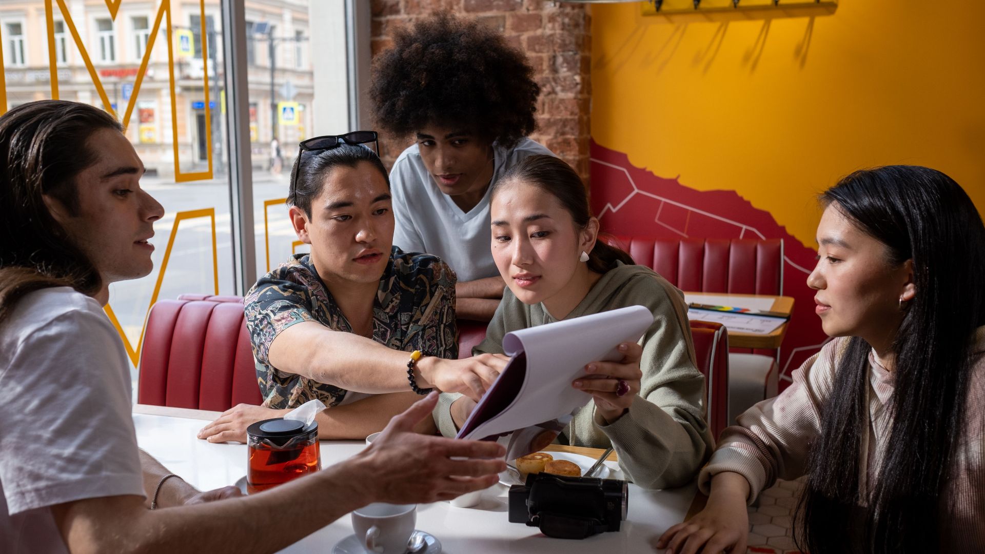 A multicultural group of young adults engaged in conversation around a table in a cafe, focused on a document.