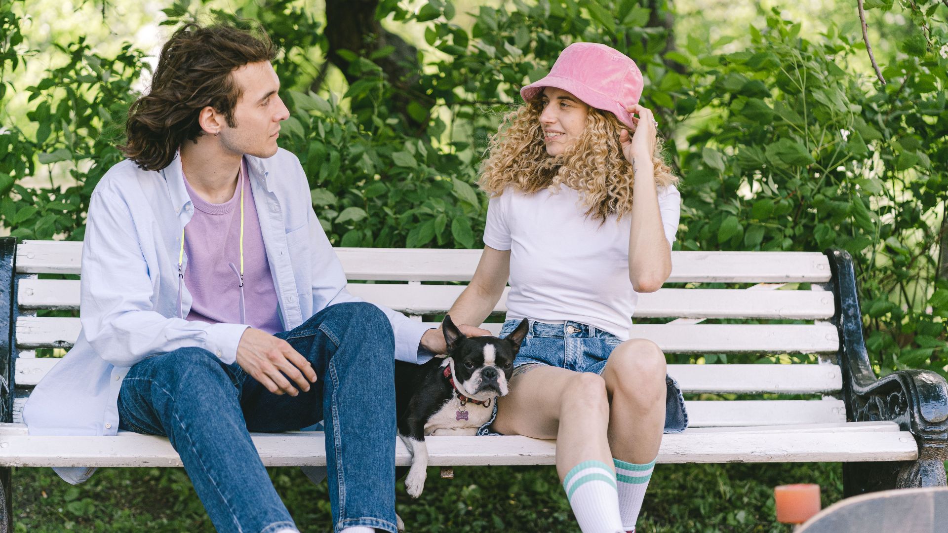 A young couple sits on a park bench, enjoying a sunny day with their Boston Terrier.