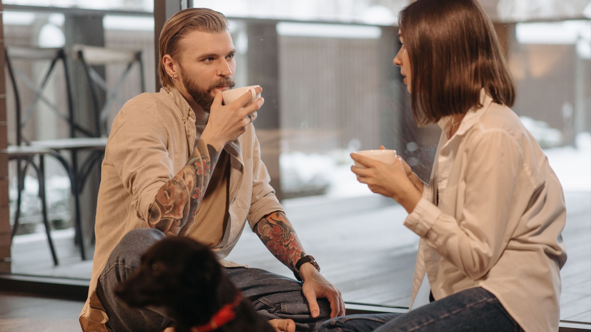 A couple sits on the floor indoors, enjoying coffee with their pet dog nearby.