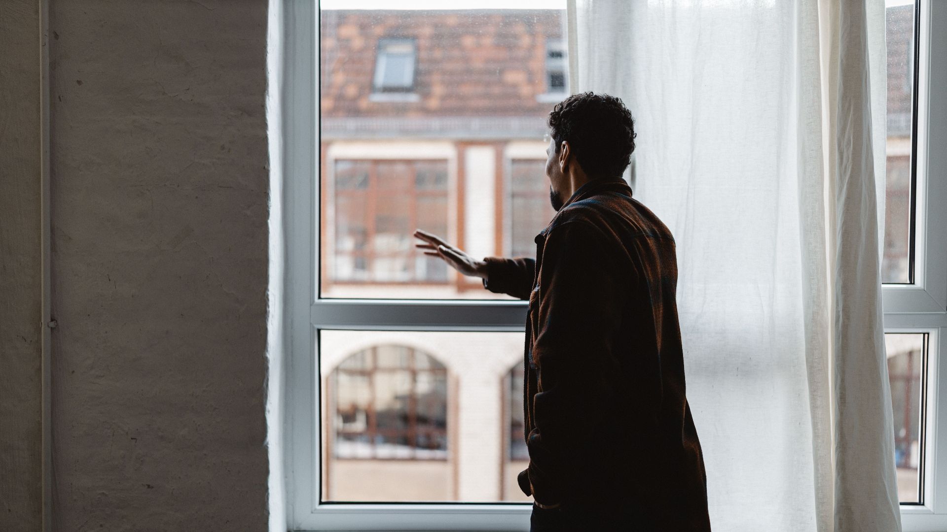 An adult gazing thoughtfully out a large window in a softly lit room.