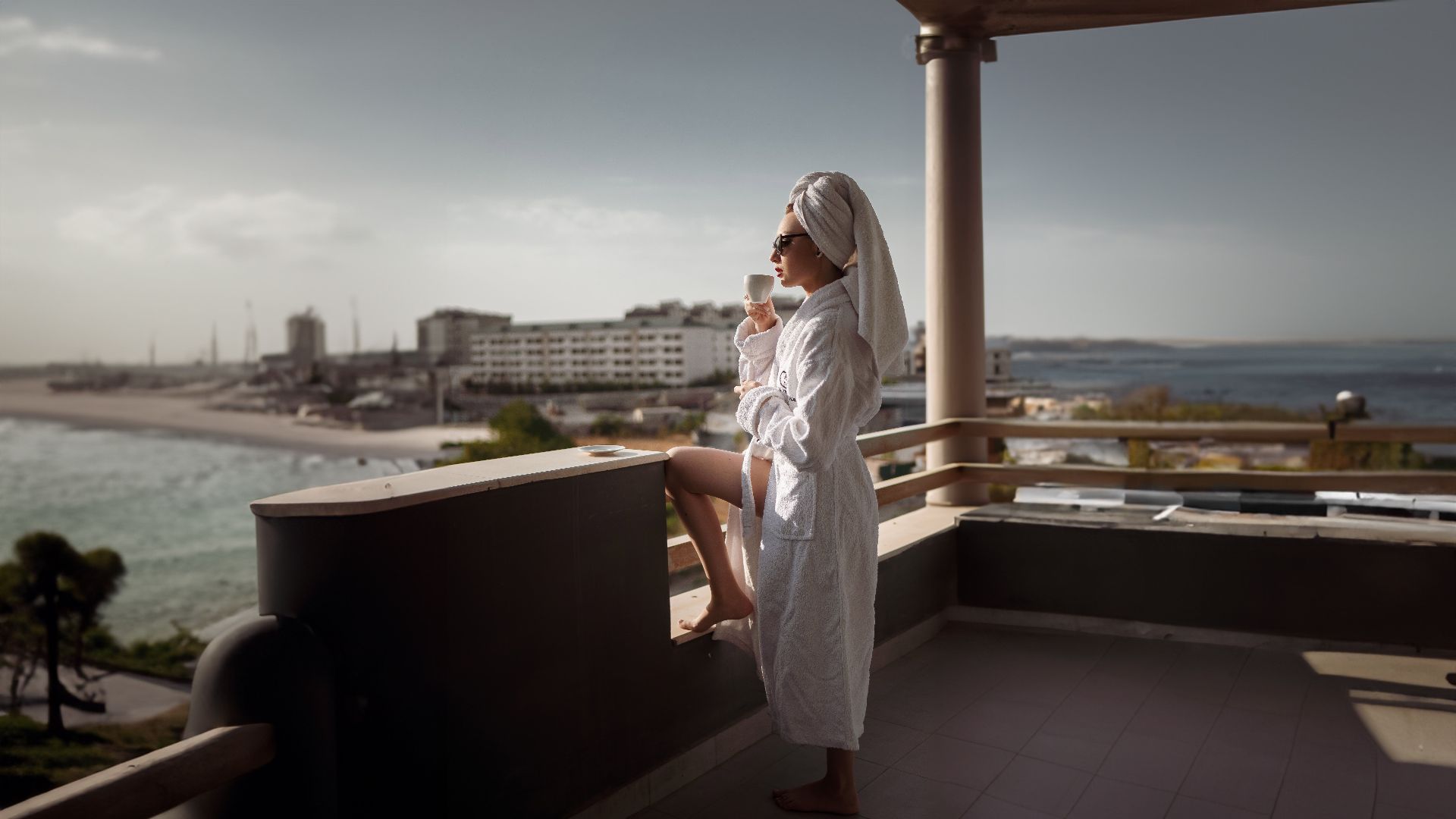 A woman in a bathrobe enjoys a drink on a sunny balcony overlooking the ocean.
