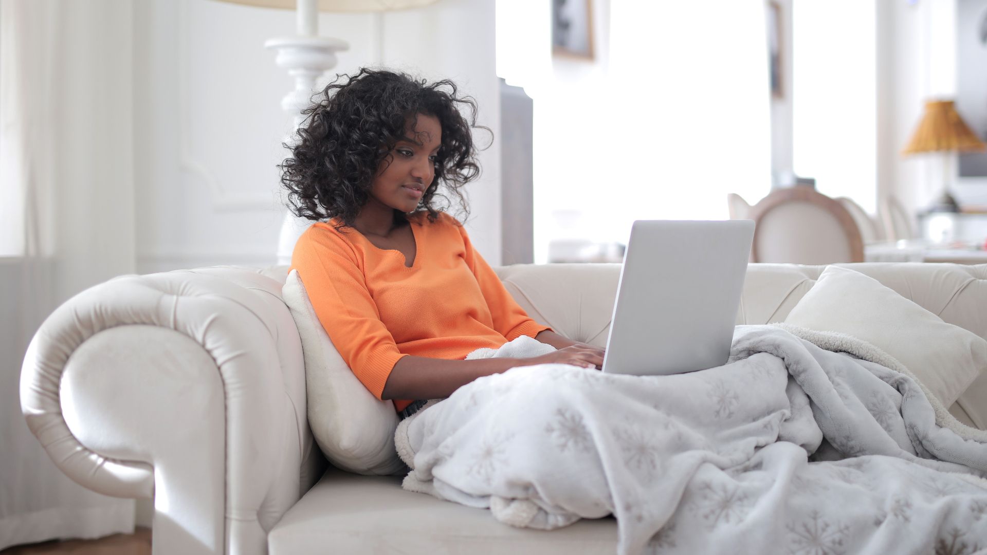 Focused African American woman sitting on sofa under blanket and browsing laptop in cozy living room