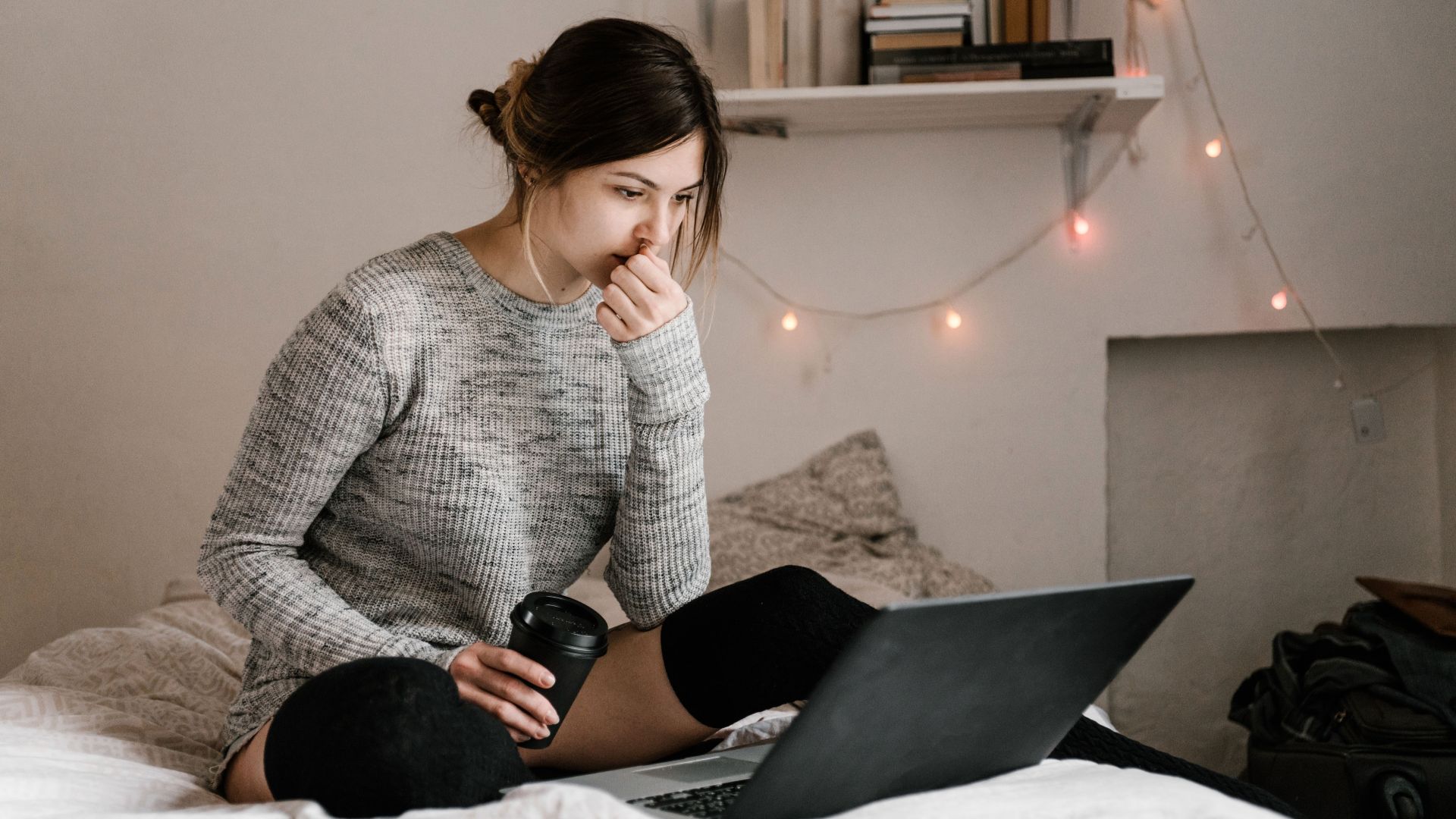 A woman in a gray sweater sits on her bed with a laptop and coffee, focused on work or study in a cozy bedroom.