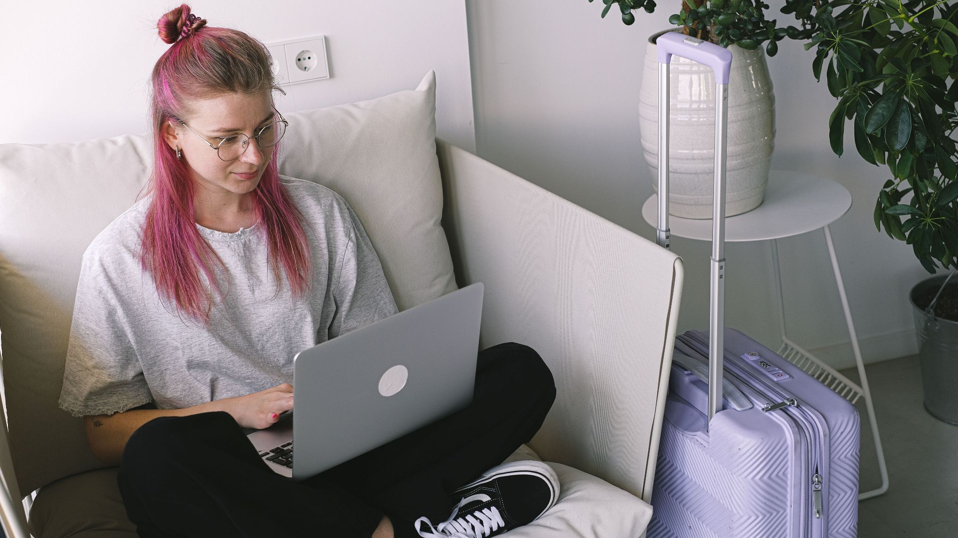 Casual young woman with pink hair works on laptop in comfortable indoor setting, travel suitcase nearby.