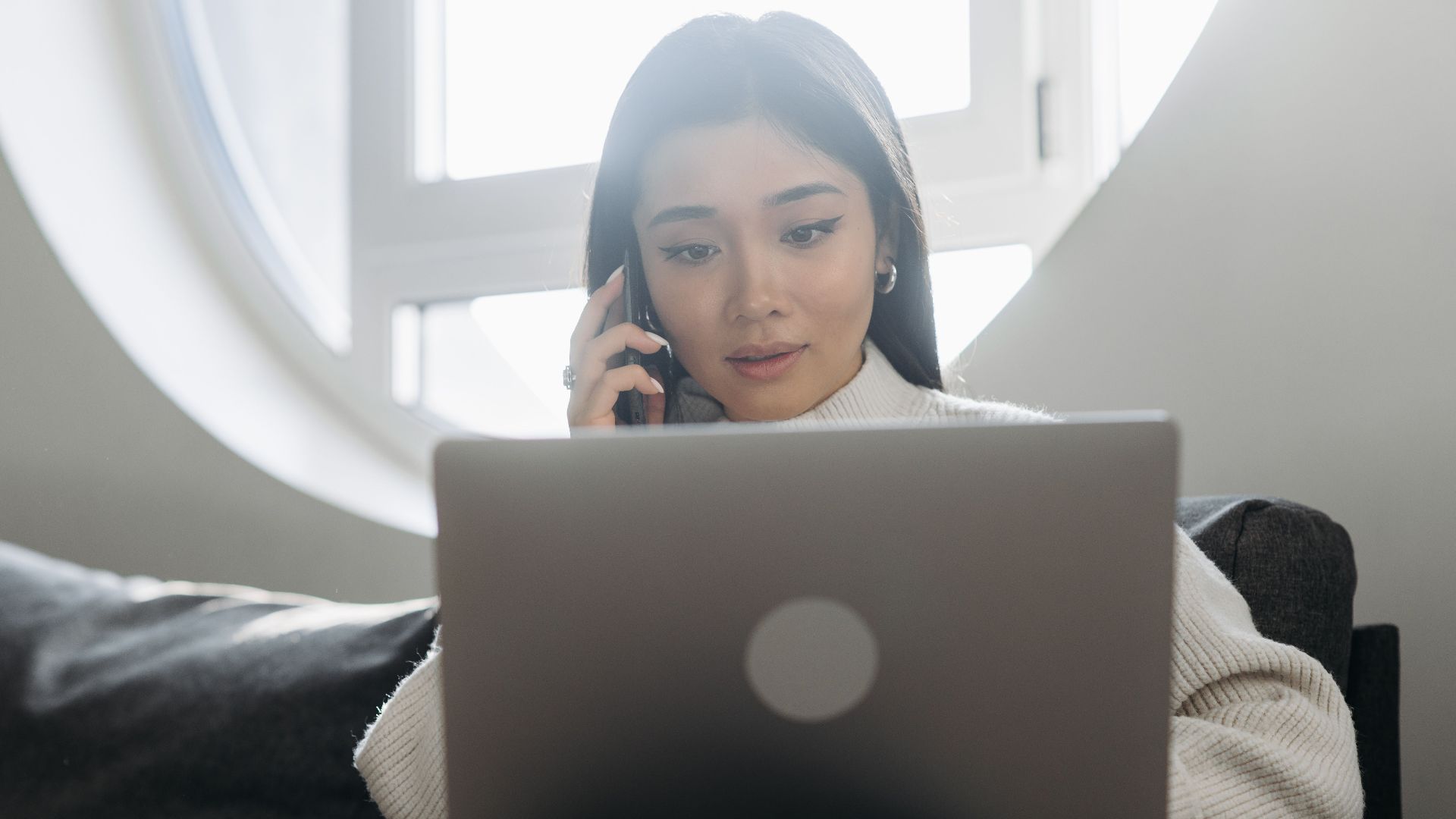 A young woman using her laptop and phone while sitting comfortably indoors.