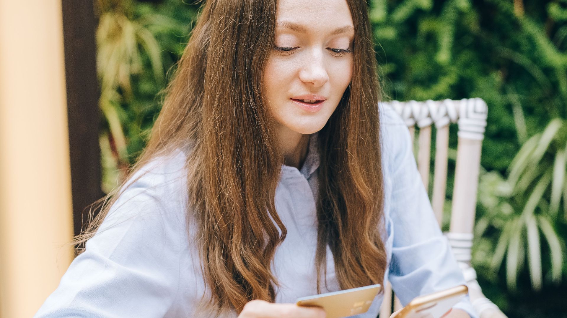 Young woman using smartphone and card for online shopping, sitting at wooden table with laptop.
