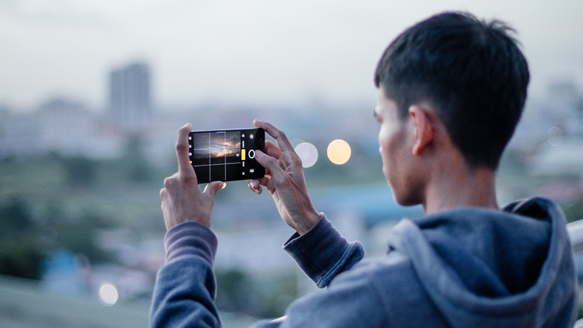 A man in a hoodie uses his smartphone to capture a cityscape from a rooftop at dusk.